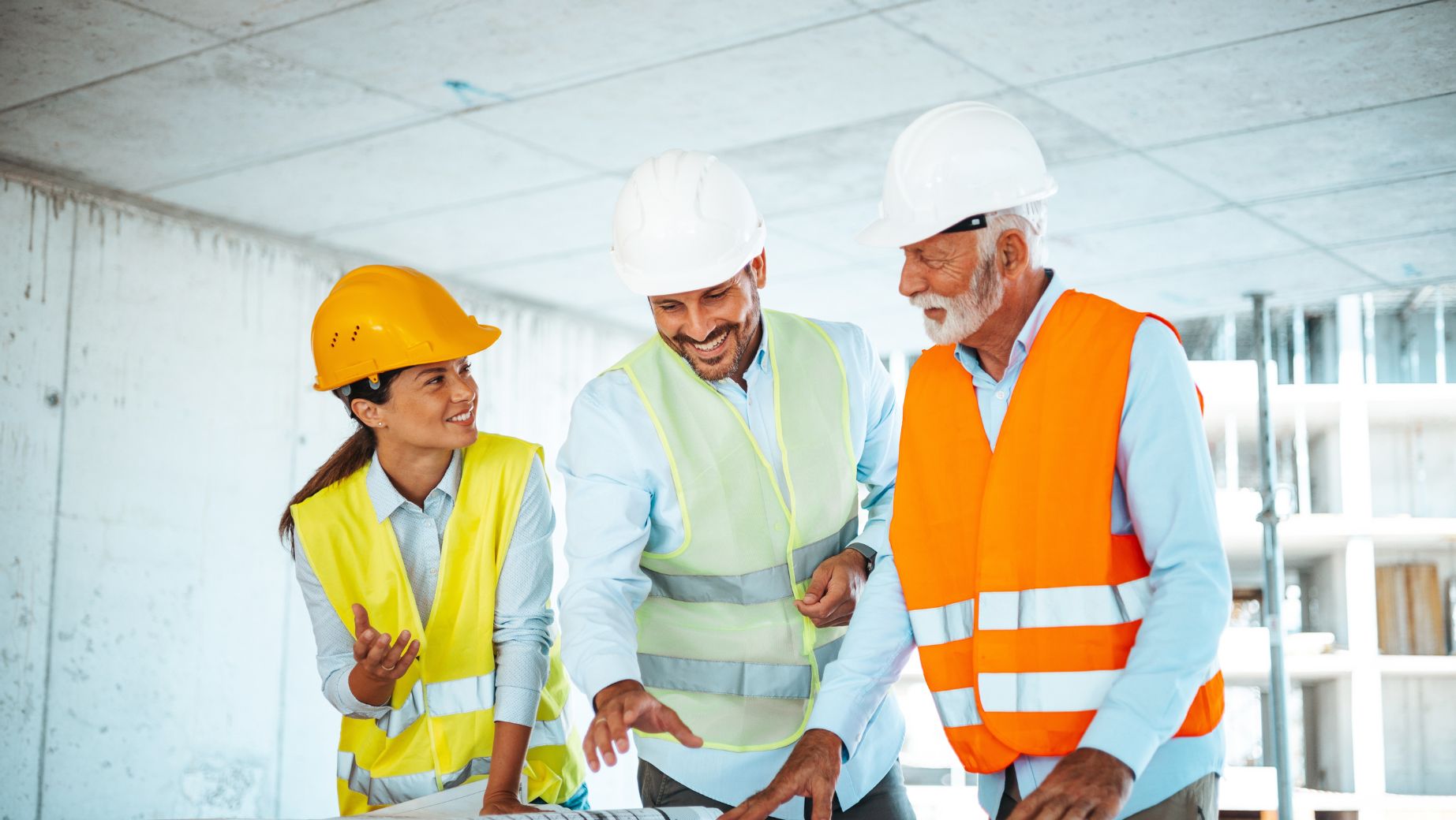 Three professionals wearing safety gear, including helmets and high-visibility vests, collaborating and reviewing plans at a construction site, illustrating teamwork in hazardous manual handling risk assessment.