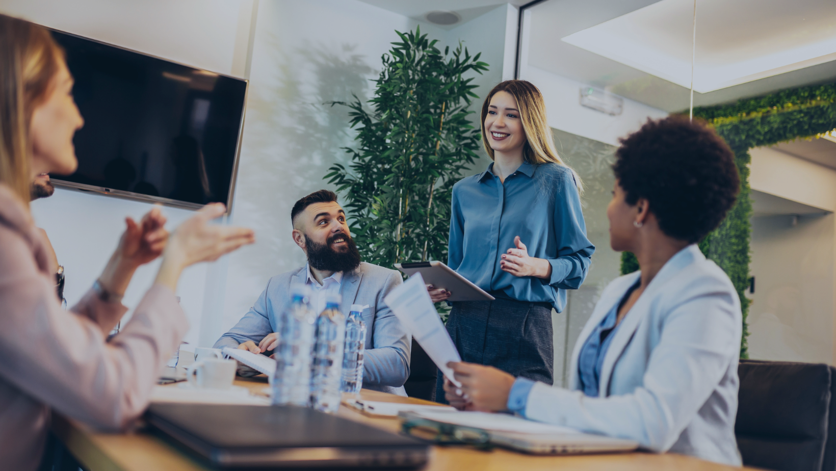 A diverse group of professionals engaged in a team discussion in a modern office, with a smiling woman standing and leading the conversation. The image represents leadership, collaboration, and the impact of people and culture transformation on workplace engagement.