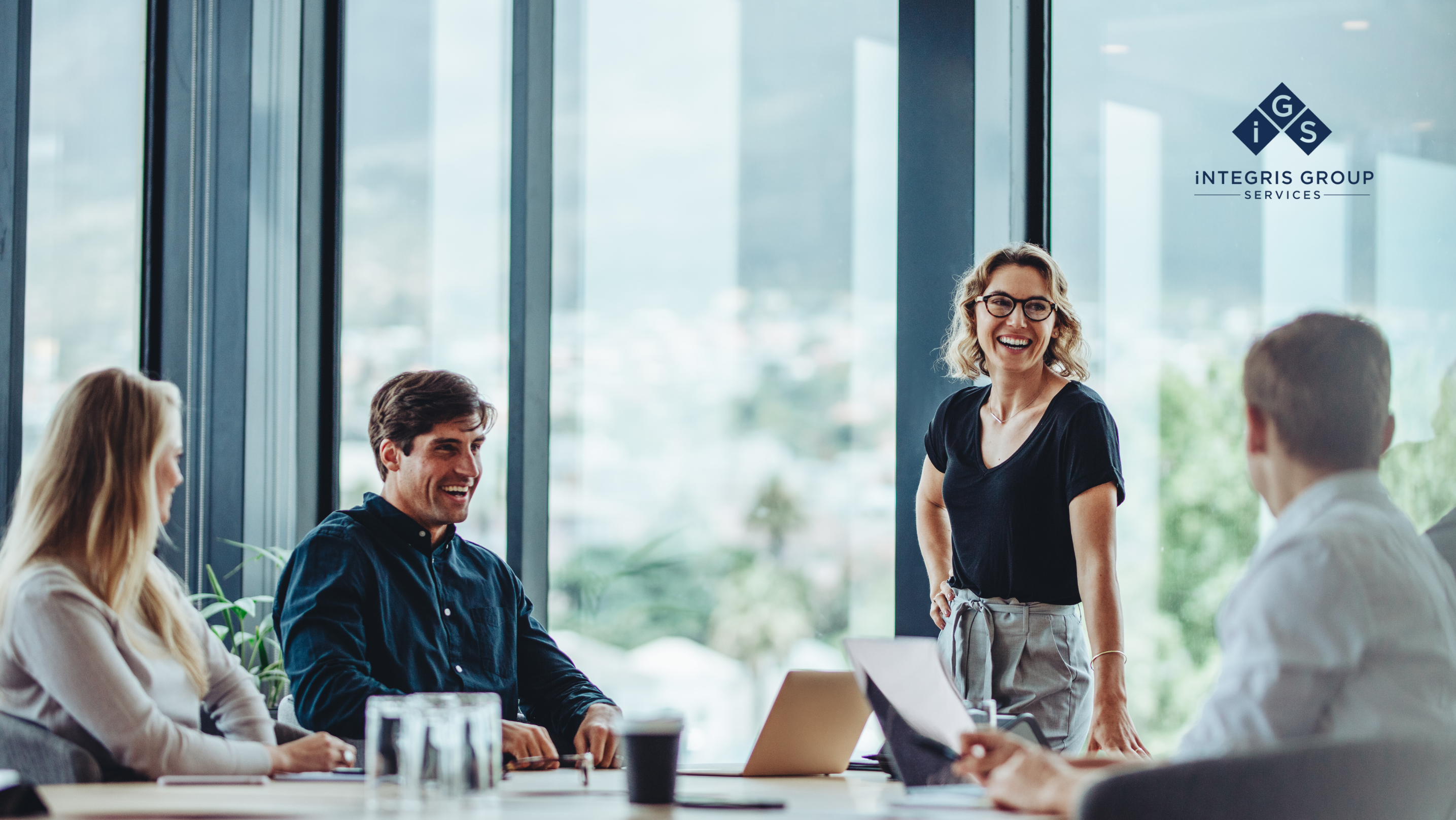 Four professionals, two men and two women, are in a meeting in a modern office at Integris Group Services. The woman standing is smiling and leading the discussion.