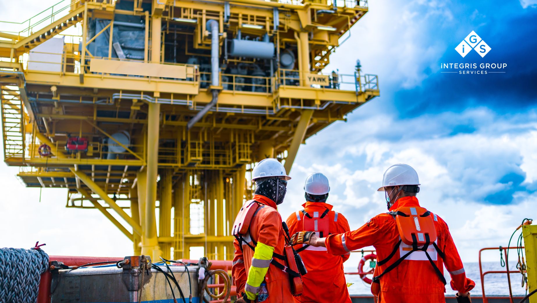 Three workers in safety gear, including helmets, high-visibility clothing, and harnesses, collaborate on a worksite near a large industrial structure, symbolising teamwork and workplace safety.