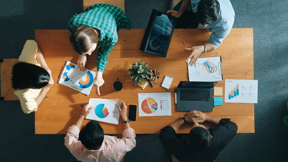 Overhead view of a diverse group of professionals engaged in a client collaboration meeting, reviewing charts and data at a conference table, highlighting teamwork and strategic planning.