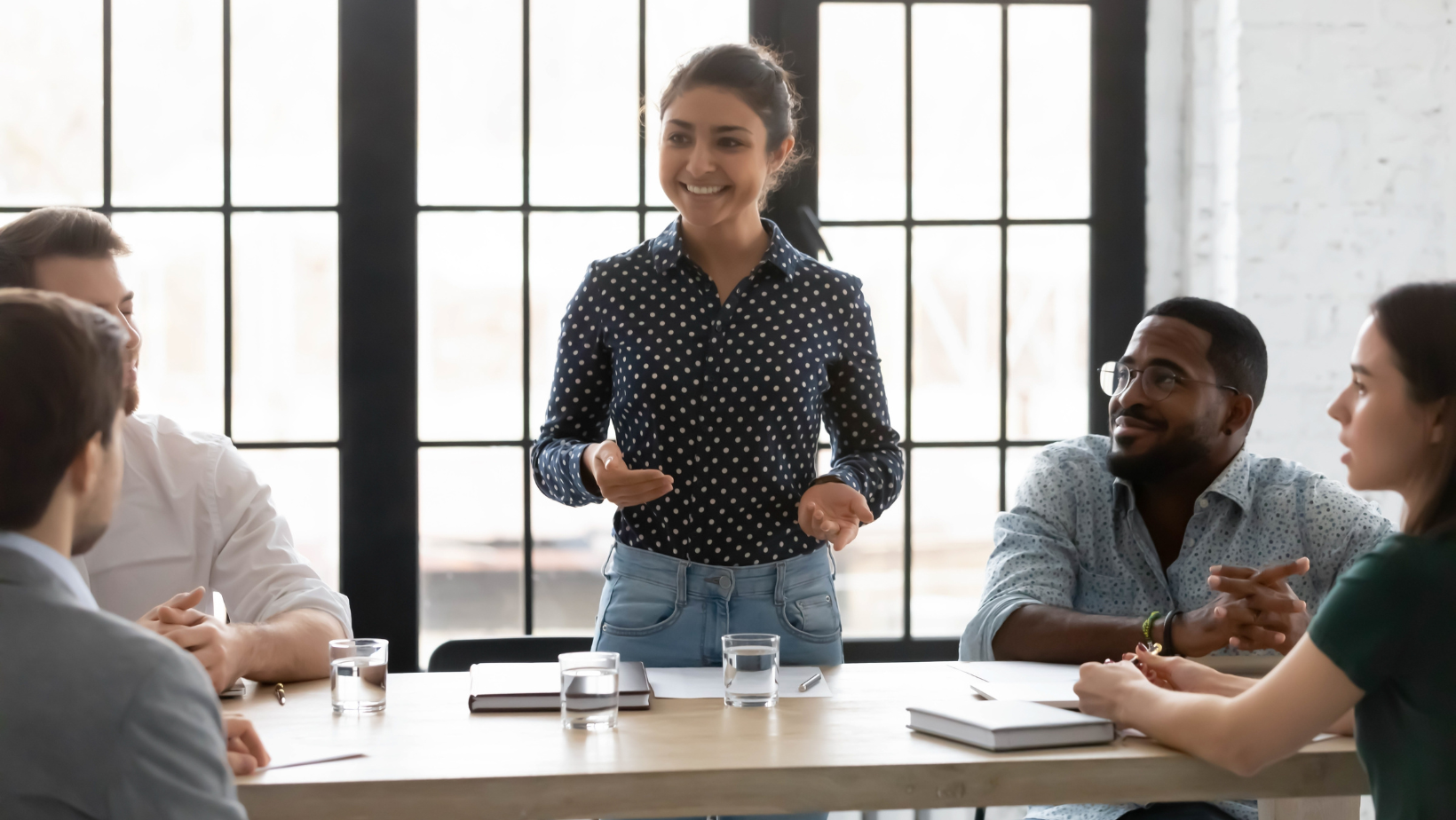 A confident woman leading a discussion in a modern office setting, engaging a diverse group of colleagues in a collaborative conversation. The image represents leadership, employee engagement, and the impact of people and culture transformation in the workplace.