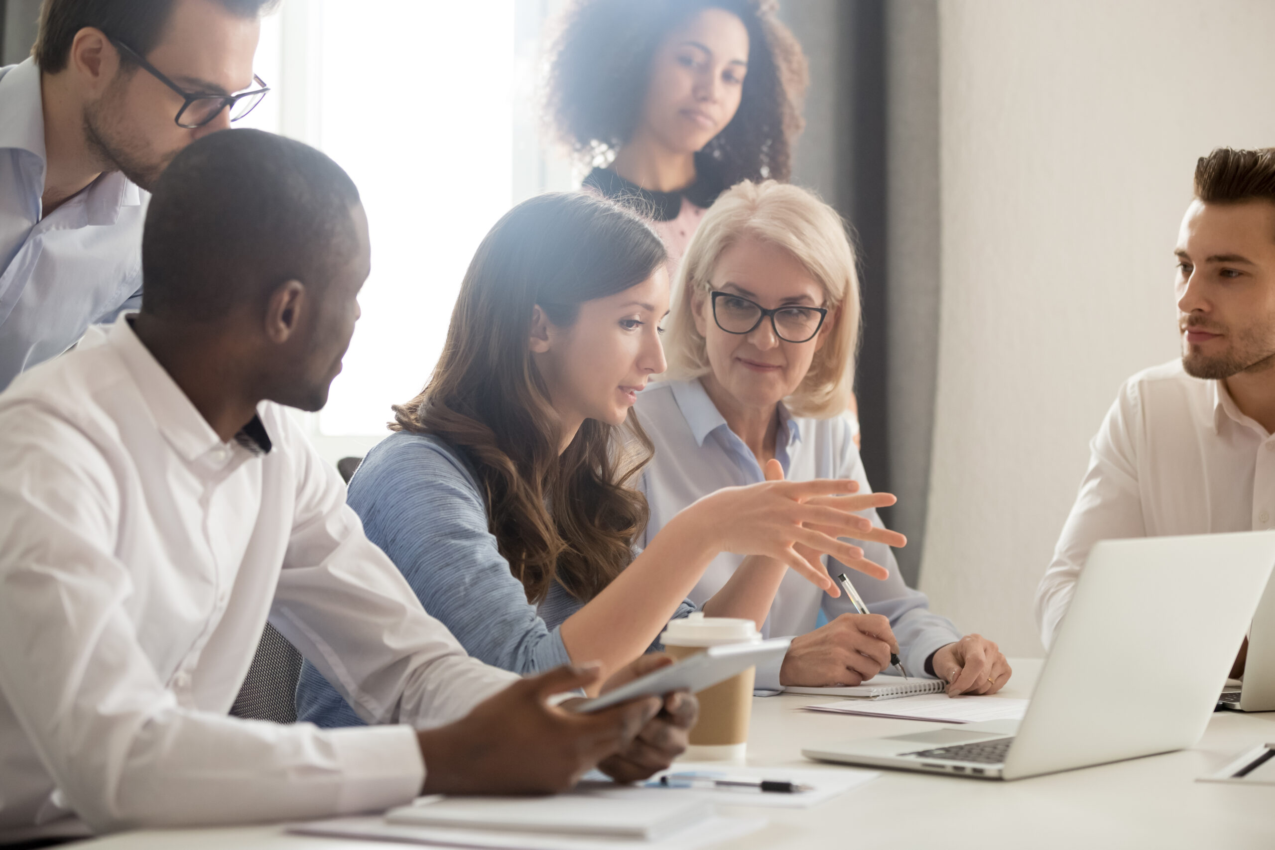 The image shows six diverse people in a meeting. A young woman speaks while others listen attentively. Laptops, notebooks, and a coffee cup are on the table, indicating a professional setting.